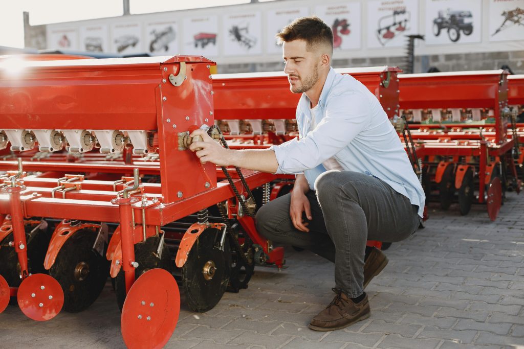 A young man examines red agricultural machinery outdoors on a sunny day.